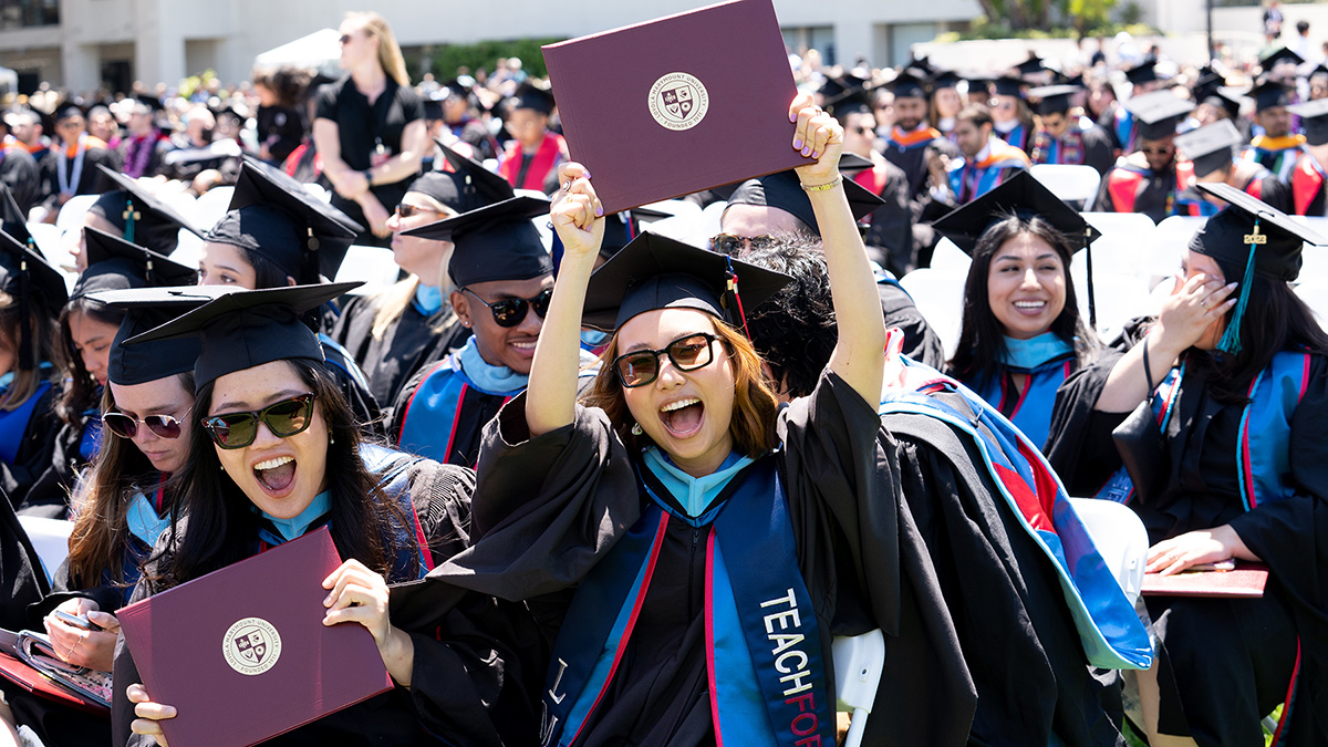 two students holding up their diplomas in front of other graduates during commencement